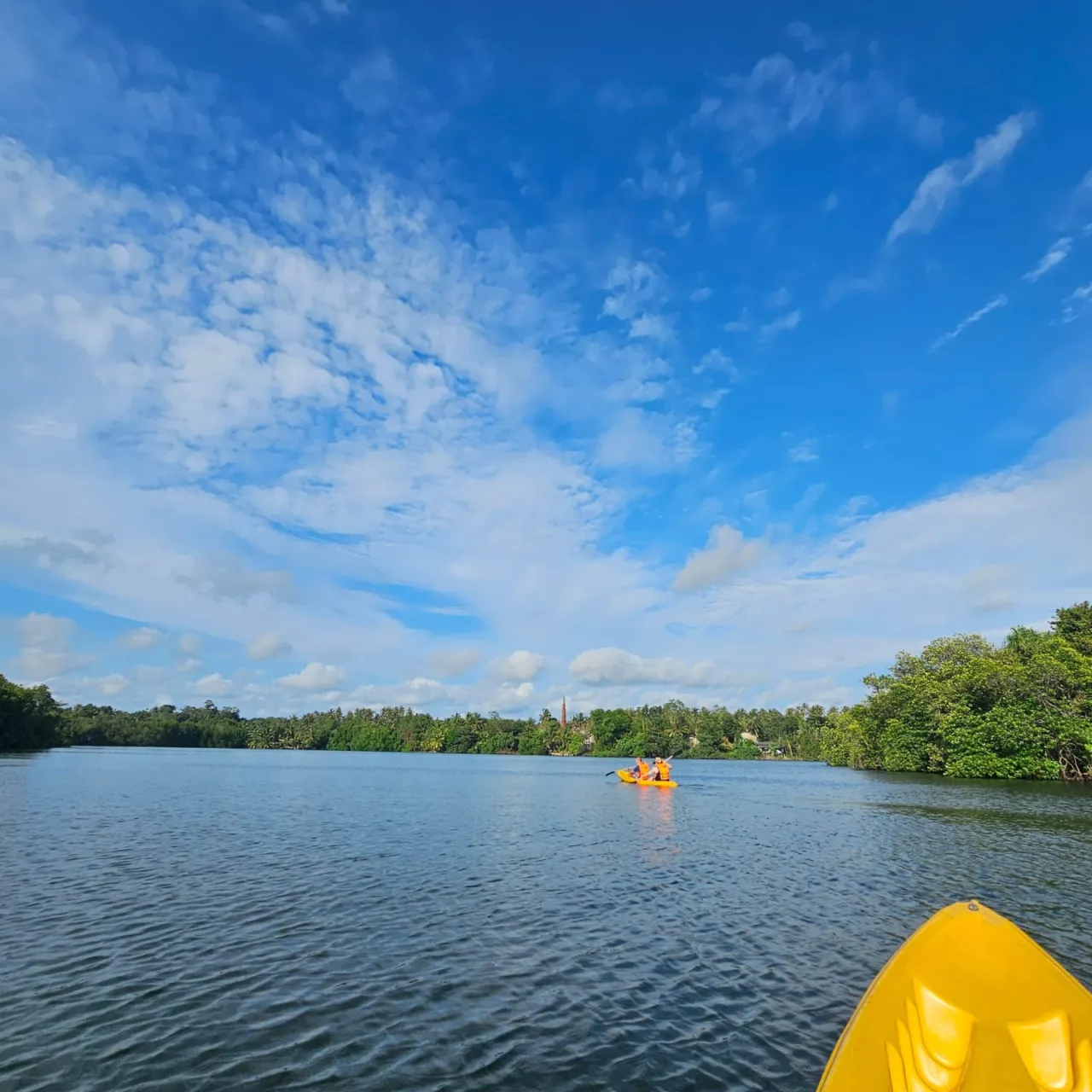 View over the bow of a yellow kayak toward distant paddlers on peaceful Rathgama Lagoon